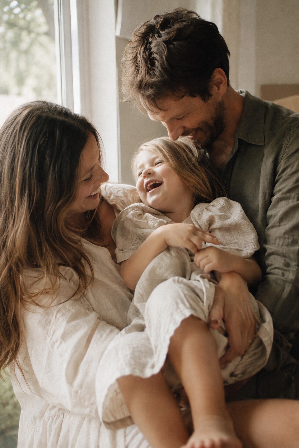 Parents and their young child laughing together beside a bright window